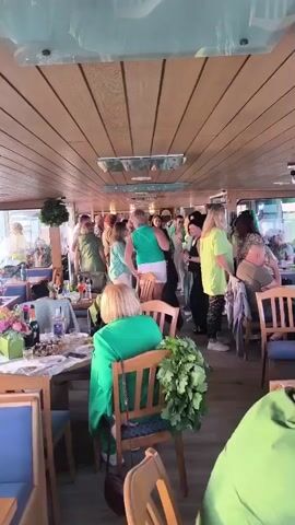 The video shows a group of people, mostly women, in a room with wooden walls and ceiling. Many of the people are wearing green clothing. Some people are standing and talking, while others are sitting at tables. There are bottles of drinks and plates of food on the tables. The atmosphere appears to be festive.