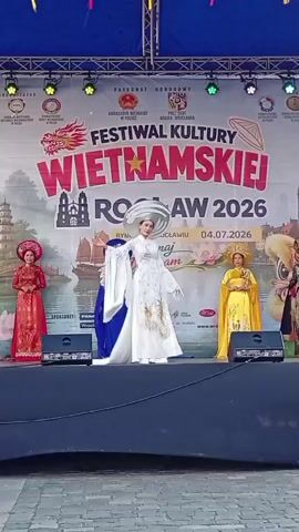 A group of women in traditional Vietnamese Ao Dai dresses are standing on a stage in front of a banner that reads "Festiwal Kultury Wietnamskiej Wroclaw 2026". The woman in the center is wearing a white dress and a large, ornate hat, and she is holding her arms out to the sides. To her left, a woman in a red dress stands with her hands clasped in front of her. To her right, a woman in a yellow dress stands with her hands clasped. The stage has speakers on either side. The background appears to be an outdoor event.