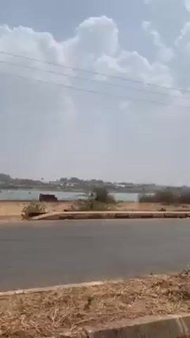 The video shows a view from a moving vehicle traveling on a road. On the other side of the road, there is a grassy, dry area with some small trees and bushes. In the distance, there appears to be a body of water with buildings lining the shore. The sky is light blue with scattered white clouds. Power lines run parallel to the road. The camera is pointed towards the left side of the car.