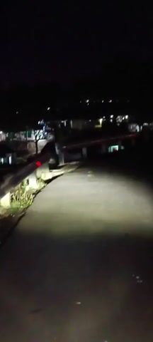 The video shows a night view of a road with a guardrail on the left side. The headlights of a vehicle illuminate the road ahead, creating a bright patch of light. In the distance, there are some lights from buildings. The road appears to be paved and curves to the right. The guardrail is made of metal and has some reflectors on it. The environment is dark, indicating it is nighttime. The camera appears to be mounted on a moving vehicle.