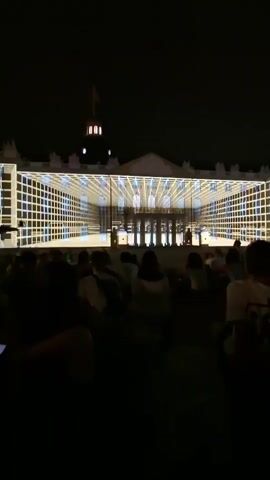The video captures an outdoor nighttime event where a crowd of silhouetted people are gathered in the foreground, facing a large building. The building is illuminated with a projected light show that creates a three-dimensional grid effect across its facade, giving the illusion of a transparent structure. The light display changes in intensity and pattern, revealing architectural details of the building beneath the projected grid. A domed structure with a flagpole is visible at the top center of the building.