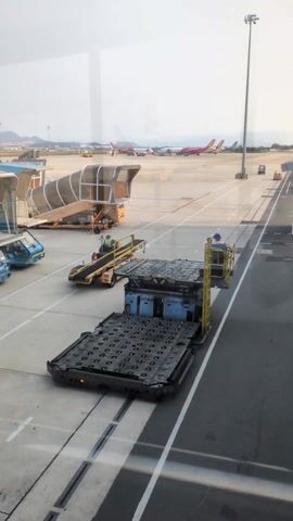 The view from inside an airport terminal shows a tarmac with several ground support vehicles. A large flatbed transport vehicle with a gridded surface and a raised platform is prominent in the foreground, occupied by a person. Another similar vehicle, possibly a conveyor belt, is behind it. Nearby, there are aircraft stairs and small blue utility vehicles.  In the distance, several commercial airplanes are parked. The light indicates it is daytime with slightly overcast weather.