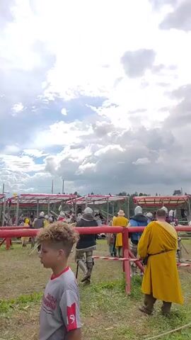 The video captures a reenactment event. In the foreground, a person wearing a red and white shirt is partially visible. The background features a jousting arena, with wooden barriers. Several individuals in historical costumes, including knights in armor and people in tunics, are present. Some are walking, while others appear to be engaged in activities within the arena. The sky is partly cloudy.