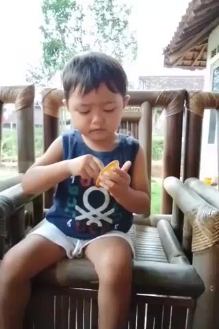 A young boy sits in a bamboo chair outdoors, wearing a dark blue sleeveless shirt with a white graphic and white shorts. He is holding a small orange object in his left hand and a small spoon in his right hand. He uses the spoon to scoop food from the orange object and brings it to his mouth. The chair has a woven seat and backrest. Behind the boy, there is a blurred background of green foliage and the roof of a building. The lighting suggests it is daytime.