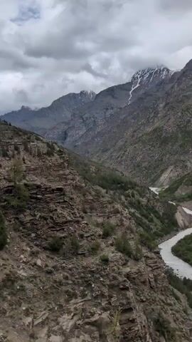 The video shows a mountain landscape. A river flows through a valley, with steep, rocky slopes on either side. The mountains are tall and rugged, with some snow visible on the peaks. The sky is cloudy, with a gray overcast. The terrain is mostly barren, with sparse vegetation growing on the slopes. There are some trees visible, as well as a few scattered pieces of litter on the ground in the foreground. The camera appears to be moving, suggesting it is mounted on a vehicle.