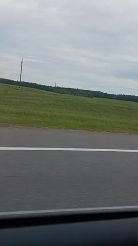A side-view of a highway with a green field and trees in the background. The sky is cloudy. A metal guardrail separates the highway from the field. There is a tall pole with what appears to be equipment on top in the distance.