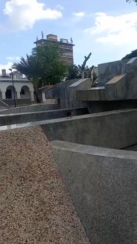 The video shows a bird, possibly a crow or raven, walking across a series of concrete structures in an outdoor setting. The bird is dark-colored and appears to be of medium size. The concrete structures are angular and appear to be part of a decorative or functional element like a fountain or a seating area. In the background, there is a tall, multi-story building and some trees. The sky is cloudy. The bird is the primary focus of the video as it navigates the various levels of the concrete structure.