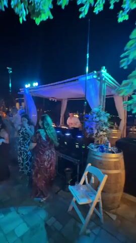 The video shows a nighttime outdoor party. In the foreground, a white chair with glasses is positioned in front of a wooden barrel. Behind the chair, a woman in a patterned dress is dancing. To her right, a DJ booth is set up under a white canopy, with a DJ operating equipment. A floral arrangement sits on the barrel. In the background, a group of people are dancing and socializing under string lights, with a building structure visible behind them. The lighting is predominantly blue and white.
