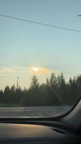 The video captures a view from inside a car, looking out through the windshield. The sun is setting, casting a warm glow and creating lens flares on the windshield. In the distance, a line of trees forms the horizon, with a cell tower visible among them. The interior of the car is partially visible, showing the dashboard and a small decorative cat figurine on top of it. The overall atmosphere is serene, suggesting a drive through a rural or natural landscape during dusk.