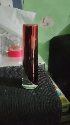The video shows a close-up, slightly shaky shot of a clear glass perfume bottle with a metallic red cap. The bottle is positioned vertically on a dark, textured surface, possibly fabric. In the background, a pale green wall is visible, with some white and grey markings on it. To the left of the bottle, a clear plastic container with a pink base is partially visible, containing small objects. The lighting is dim, casting shadows and highlighting the reflective surface of the cap.