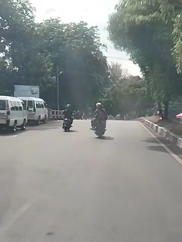 The video shows two people riding motorcycles on a paved road. The road is lined with trees and a concrete barrier. There is a white van parked on the left side of the road. The sky is overcast. The video is taken from a moving vehicle.