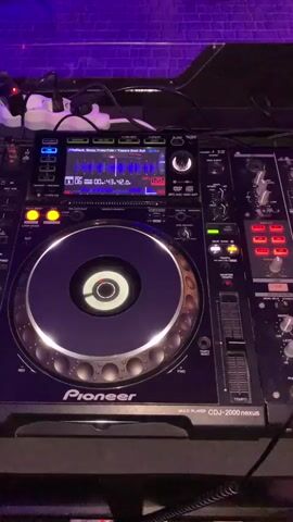 The video shows a close-up view of a DJ setup in a dimly lit room, likely a club or bar, with purple lighting visible in the background. The primary focus is on a Pioneer CDJ-2000 Nexus multi-player and a mixer. The CDJ has its jog wheel illuminated and is displaying song information on its screen, indicating a track is playing. Various buttons and knobs on the DJ equipment are visible, some lit up. To the right of the CDJ is a mixer with multiple channels, sliders, knobs, and illuminated buttons, suggesting it's in use. Part of a person's legs and feet, wearing white sneakers, are visible at the bottom right of the frame, indicating someone is operating the equipment. The overall atmosphere appears to be that of a DJ preparing to or actively performing.
