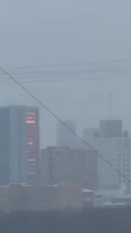The video shows a cityscape on a very overcast day. There are several buildings visible, some of which appear to be residential buildings and one large white building, perhaps an office building. There are power lines stretching across the scene, partially obstructing the view. One building in the distance has a reddish glow reflecting off of its windows. The foreground is obscured by a dark mass, likely trees or another building in the immediate vicinity of the camera. Snow covers the ground and rooftops. The overall atmosphere is muted and hazy due to the weather conditions.