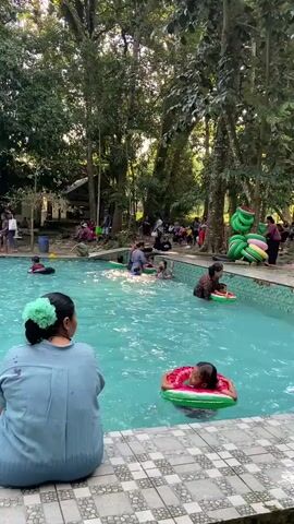 A swimming pool is surrounded by trees and a tiled edge. Several people are in the pool, some of whom are children. Many of the children are using inflatable rings for buoyancy. One child in the foreground is wearing a watermelon-shaped ring. Other people are sitting or standing around the pool. There are also several inflatable rings stacked on the right side of the image. The overall scene depicts a recreational swimming area.