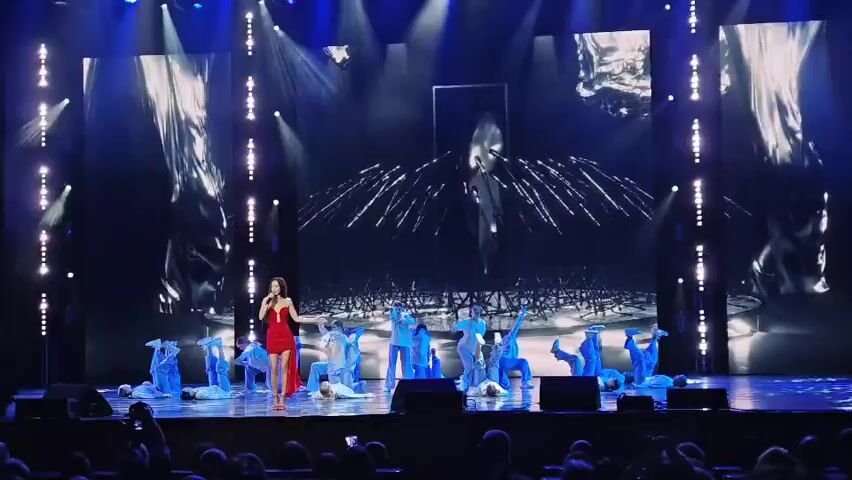 A female singer in a red dress stands on a stage, holding a microphone and singing. She is surrounded by dancers dressed in blue, some of whom are lying on the stage while others are in various poses of movement. The stage features a large screen backdrop displaying abstract, reflective, and geometric patterns with streaks of light resembling fireworks. The lighting is dramatic, with spotlights illuminating the stage. The audience is visible in the foreground, seated in the darkness.