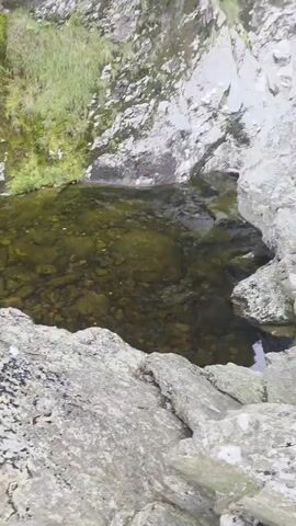 The video shows a close-up, slow-motion view of a waterfall cascading down a rocky, moss-covered cliff face. The water is clear and appears to be flowing over rough, gray rocks. Green moss and grass grow in patches on the rocks. At the bottom of the waterfall, there is a pool of clear water with rocks visible beneath the surface. The perspective is from slightly above and to the side of the waterfall, looking upwards at the falling water and the cliff face. The lighting is natural and diffused, suggesting an overcast day.