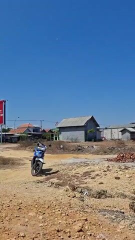 The video begins with a wide shot of a coastal area under a clear blue sky. In the background, there are several buildings, some appearing to be warehouses or industrial structures, and a smaller, simpler building with a gray roof. The foreground consists of a rough, rocky terrain with some loose bricks and scattered debris.

The camera then pans to the right, transitioning to a closer view of a young man sitting on a rocky embankment by the sea. He is wearing a gray hooded jacket over a purple shirt and a dark vest. He has short, dark hair and a mustache. His left leg is crossed over his right, and he is looking out towards the sea. A black motorcycle helmet rests on the rocks next to him.

The background shows the turquoise sea, with a wooden pier extending into the water. Small structures or houses are visible on the pier. The water is calm, with gentle waves lapping against the shore. The video concludes with a further pan to the right, focusing on the vast expanse of the sea and the distant pier under the bright, cloudless sky.