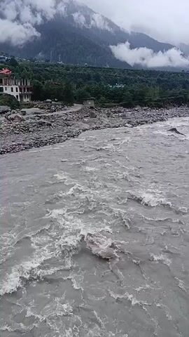 A wide, muddy river flows through a valley. On the left bank, there is a multi-story building with a white facade and red accents, situated on a rocky slope dotted with trees. Further back, more buildings and dense green foliage cover the hillside. In the background, towering mountains are partially obscured by thick, grey clouds. The river itself is turbulent, with white foam and churning water indicating a strong current. The scene is overcast, suggesting a cloudy or possibly rainy day. The video appears to be shot from a bridge or elevated position overlooking the river and the surrounding landscape. There are no visible people or animals in the frame.