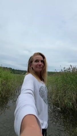 A woman with blonde hair, wearing a white sweatshirt with a circular design, is holding a phone out as if taking a selfie. She is standing in shallow water with a rocky bottom. Tall reeds line the edge of the water and stretch into the background. The sky is overcast. The camera pans to the left and then to the right, showing more of the reeds and the lake.