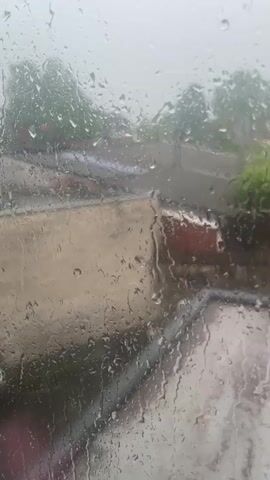 The video shows a view through a window on a rainy day. Raindrops cover the glass, obscuring the scene outside, which includes a road, a concrete patio, some greenery, and what appear to be some buildings or structures in the distance. The atmosphere is gray and overcast. No specific people or actions are clearly visible.