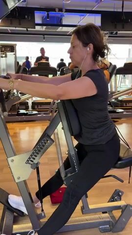 A woman is using a seated row machine at a gym. She is wearing a black t-shirt, black leggings, and white sneakers. She has earphones in and is pulling the handles of the machine towards her chest. The machine is silver and has a back pad for support. Other people can be seen working out in the background on treadmills and other exercise equipment. The gym has a wood floor and windows overlooking an outdoor area.