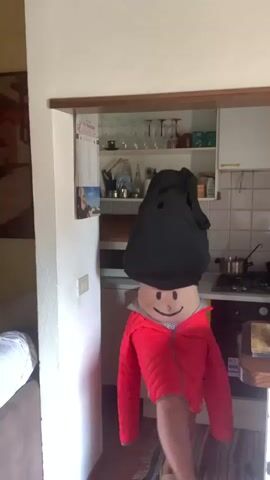 This media file shows a person in a kitchen wearing a black hooded garment over their head, obscuring their face. A red quilted jacket is worn over their torso, and a smiley face is drawn on their stomach. Their legs are visible below the jacket. The person appears to be moving, with their body in various poses, suggesting dancing or playful movement.

The setting is a kitchen, with white cabinets, a tiled backsplash, and a stovetop visible in the background. Shelves above the counter hold various kitchen items, including glasses and jars. A calendar with Italian text is hanging on the wall to the left. The lighting suggests it is daytime. The overall mood is lighthearted and whimsical. The location is San Quirico d'Orcia, Italy, which can be inferred from external information.