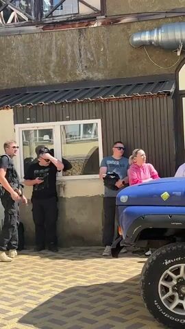 The video shows a group of people gathered around several off-road vehicles, which appear to be UAZ jeeps. The vehicles are parked in an outdoor area with a building in the background. The people are dressed casually and some are interacting with each other and the vehicles. One person is opening the door of a blue UAZ jeep, while others stand nearby. The environment suggests a sunny day. The video cuts between different scenes of the jeeps and people.