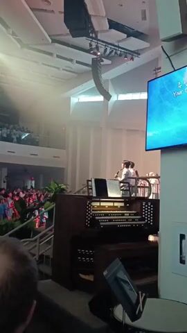The video shows a large gathering of people, likely in a place of worship, with a stage area in the foreground. An organ is visible in the lower right corner of the frame. On the stage, several individuals are present, some holding microphones and appearing to lead the congregation. The audience, composed of many people, is arranged on multiple levels, with some standing and others seated. Bright spotlights illuminate the stage and parts of the audience. A large screen is mounted on the wall in the background, displaying what appears to be text. The overall environment suggests a performance or service.