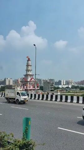 The video shows a multi-tiered temple structure with a spire on top, painted in white and pink, with red and orange accents. It is situated on the side of a road, with a highway barrier in the foreground. A yellow backhoe loader is visible parked on the side of the road. Buildings and trees are in the background. The sky is partly cloudy. The camera appears to be moving, likely from a vehicle.
