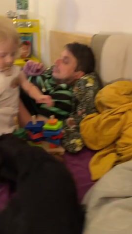A man is asleep on a couch, leaning on his side. A young child is sitting next to him, playing with a stacking toy. A black dog is lying on the couch in front of them. The room appears to be a living room or bedroom, with a couch, pillows, and a blanket visible.