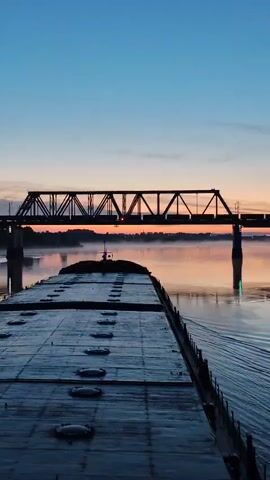 The video shows the rear deck of a barge moving on water towards a bridge, likely at dawn or dusk. The sky transitions from a deep blue to a pale orange. The water reflects the colors of the sky, and there is a light fog on the water's surface. The barge is gray and features a number of round hatches. The bridge has a dark, metal truss design and is also reflected in the water.
