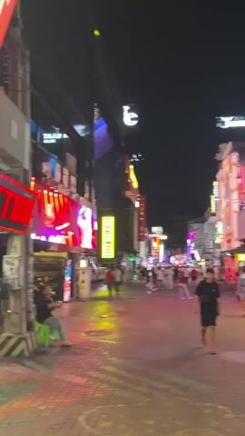 The video shows a street scene at night, likely a tourist area. The street is paved with cobblestones and illuminated by neon signs and lights from businesses lining both sides. There are several people walking along the street, some are looking at their phones, others are walking in groups. One person is sitting on a small green stool on the left side of the street. Buildings on either side have illuminated signs, some with text that is difficult to read due to the lighting. The overall atmosphere is lively and urban.