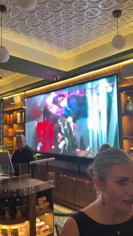 Inside a well-lit restaurant, several TV screens display the same video of a woman in a winter setting. The restaurant is filled with patrons, and staff members can be seen interacting with customers. The interior design features a patterned ceiling with spherical hanging lights and green accents. There are shelves stocked with products, and a counter area where a staff member is working. People are walking around and socializing within the restaurant's dining area.