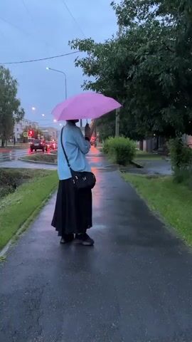 A person holding a pink umbrella walks on a wet sidewalk on a cloudy day. The person is wearing a light blue shirt over a black dress and a black crossbody bag. The sidewalk is next to a grassy area and a street with cars and buildings in the background. The sky is overcast, suggesting it is either early morning or late evening, and it appears to be raining or has recently rained, as the sidewalk is wet and reflective. The person is walking away from the camera, and the umbrella is tilted, as if protecting them from the rain. The scene appears to be in a residential or urban area. There is a traffic light visible in the distance with green and red lights. No text is visible in the media.