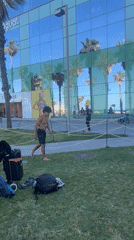 Men practice backflips on Barcelona grass near mirrored building