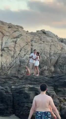 The video shows a rocky beach landscape. In the foreground, a shirtless man wearing patterned swim trunks stands with his back to the camera. On a higher rock formation in the middle ground, a couple is embracing. The man in the couple is wearing a white shirt and khaki pants, holding a red can, while the woman wears a white dress. Behind and to the left of them, another man stands shirtless in red swim trunks. The backdrop features a large, light-colored rocky cliff beneath a cloudy sky.