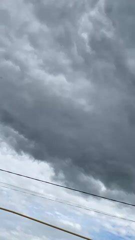 A vertical video shows a cloudy sky with some lighter patches and blue sky visible in the distance. There are power lines in the foreground, one running horizontally across the middle of the frame, and another closer to the bottom. The clouds are dark gray and appear to be moving.