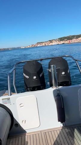 The view from the stern of a white motorboat showcases two black-covered Mercury outboard motors. The boat cruises across a body of water, creating a wake behind it. In the distance, a town sprawls along a coastline under a clear blue sky. The boat's wooden deck, stainless steel railings, and a gray bumper are visible.