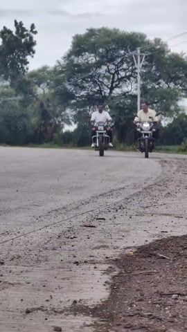 Two men ride motorcycles down a paved road. The man on the left is wearing a white shirt and dark pants. The man on the right is wearing a light-colored shirt and dark pants. They are both looking at the camera. In the background, there are trees and a utility pole. The sky is overcast. The road appears to be dusty and worn, with tire tracks visible.