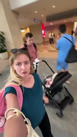 A woman with blonde hair and sunglasses on her head, wearing a teal shirt and a pink backpack, is walking forward while holding a camera. She is pushing a black stroller with a baby inside. Behind her, a man in a pink t-shirt and another man in a blue shirt are walking. The environment appears to be an indoor public space, possibly an airport or a shopping mall, with tiled floors and a curved ceiling. In the background, there are other people, escalators, signs, and displays. The video is shaky and appears to be a first-person perspective recording.