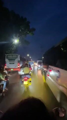 A first-person perspective video captures a person riding a motorbike on a busy street at night. The road is wet, reflecting the bright headlights of vehicles. Various cars, buses, and motorbikes are visible, with some vehicles' taillights glowing red. Streetlights illuminate the path ahead. The sky is dark blue, with trees visible on the left side of the road.