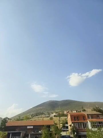 The video pans across a sky with a few clouds, revealing a town nestled at the base of a large hill. Buildings, some with red-tiled roofs, are visible, along with trees and vegetation. A white car is parked on the right side of the frame.