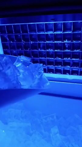 This is a close-up, first-person perspective video of an ice dispenser. Ice cubes are being dispensed into a container. The ice is clear and some of it is melting. The background is a grid of ice cube molds, illuminated by blue light. The dispenser appears to be an automatic ice maker.