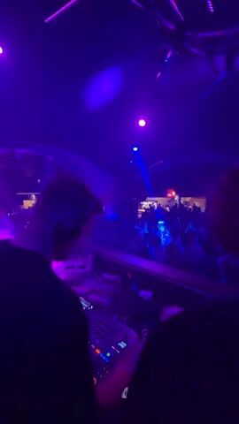 The video shows a DJ in a nightclub or outdoor event. The DJ, wearing headphones and a dark shirt, is operating a DJ mixer and turntables. The background shows a crowd of people, illuminated by purple and blue lights, as well as laser beams. The event appears to be energetic, with people moving and dancing. Some structures, possibly buildings or canopies, are visible in the background, giving the impression of a large outdoor gathering.