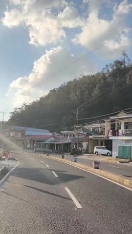 The video pans across a roadside scene with a gentle curve in the asphalt road. On the left, a row of buildings appears to be shops or houses, some with awnings and signs, with a few Indonesian flags visible. A white van is parked on the right side of the road. The sun is shining brightly, casting lens flare and illuminating the sky, which is dotted with white clouds. A forested hill rises in the background. Streetlights and power lines are present along the road.