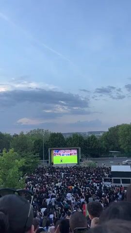 A large crowd of people has gathered outdoors in front of a stage. The crowd is predominantly dark-haired and the people are dressed in casual clothing. Many are holding up their phones as if taking pictures or videos. In the background, there is a large screen displaying text. Beyond the crowd and stage, there is a tree line and distant city buildings. The sky is partly cloudy, with a mix of blue and white and grey.
