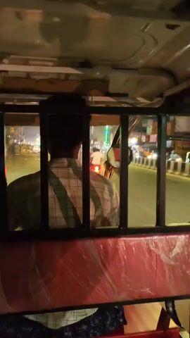 The video begins from the inside of an auto-rickshaw, looking through a barred window. A person, seen from behind, is driving the auto. The driver is wearing a checkered shirt. Outside the auto, the street is illuminated with lights, suggesting it is nighttime. Vehicles and buildings are visible, with some decorations or lights on them. The auto then passes a colorful food truck adorned with lights and pictures of food items like ice cream and drinks. The truck has text in Hindi. The video then shows the side of the truck, which is also decorated with various colorful designs and cartoon-like images. A person is standing near the front of the truck. The environment appears to be a street or roadside area at night.