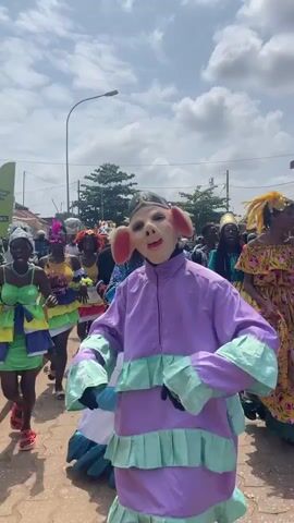 A group of people in costumes are walking down a street. In the foreground, a person is wearing a pig mask and a purple and mint green costume. To their left, a woman in a yellow and green costume is dancing. Behind them, other people are walking in colorful outfits, some with masks or elaborate headpieces. The sky is cloudy.