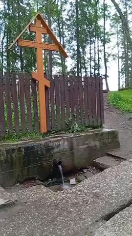The video shows a natural spring in a wooded area. A tall, wooden Orthodox cross stands behind a rustic wooden fence. The spring water flows from a dark pipe into a concrete basin, creating a small waterfall. The setting appears to be a natural, outdoor location, likely in a forest, given the trees in the background. The overall atmosphere seems peaceful and serene, possibly a place of religious significance or quiet reflection. There is no visible indication of the time of day or specific weather conditions, though the lighting suggests it might be daytime. The location cues, such as the Orthodox cross, point to Novoilinsky, Russia, as indicated. No people are visible in the video.