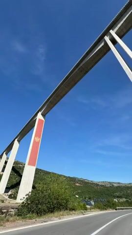 The video captures a low-angle view from a vehicle driving along a winding road beneath a large concrete bridge. The bridge features multiple tall, slender pillars, some of which have red and gold decorative elements. The sky is clear and blue with scattered white clouds, and green, hilly terrain with some sparse structures is visible in the background. The vehicle moves forward, passing by various sections of the bridge and its supports.